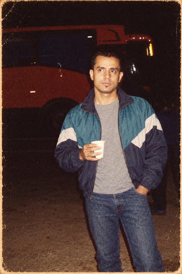 Mahabir in 2008, styled to look like a faded 1980s photo: a windbreaker and jeans, holding a paper cup at a truck stop at night.